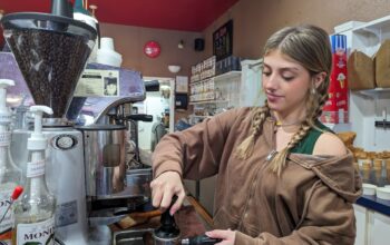 A woman with blonde hair and a brown sweatshirt pulls shots for an Americano at a silver espresso machine.