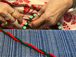 In the foreground of the picture is a blue tablecloth with red and green yarn strewn across it. It is an up-close picture of a woman's hands. In her hands, she holds a green loom and red and green yarn.