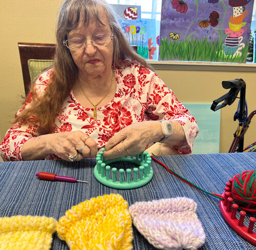 A woman with long gray hair and a white blouse with red flowers on it sits at a table with a blue tablecloth. She has a turquoise loom in her hands with red and green yarn tied to it. To the side, there is a red loom and a blue loom. In front of her are three completed hats. Two are yellow and one is purple.