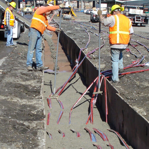 Three workers in orange vests and blue jeans stand on either side of a gray narrow trench. The two workers in the foreground of the picture are holding gray poles. Red and blue caution tape is draped into the trench. The trench in the foreground has gray cement in it. The third worker in the background is standing next to the spout on a cement truck. Gray cement is pouring down the spout into the trench.