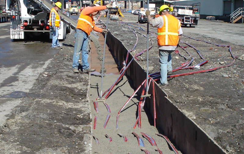 Three workers in orange vests and blue jeans stand on either side of a gray narrow trench. The two workers in the foreground of the picture are holding gray poles. Red and blue caution tape is draped into the trench. The trench in the foreground has gray cement in it. The third worker in the background is standing next to the spout on a cement truck. Gray cement is pouring down the spout into the trench.