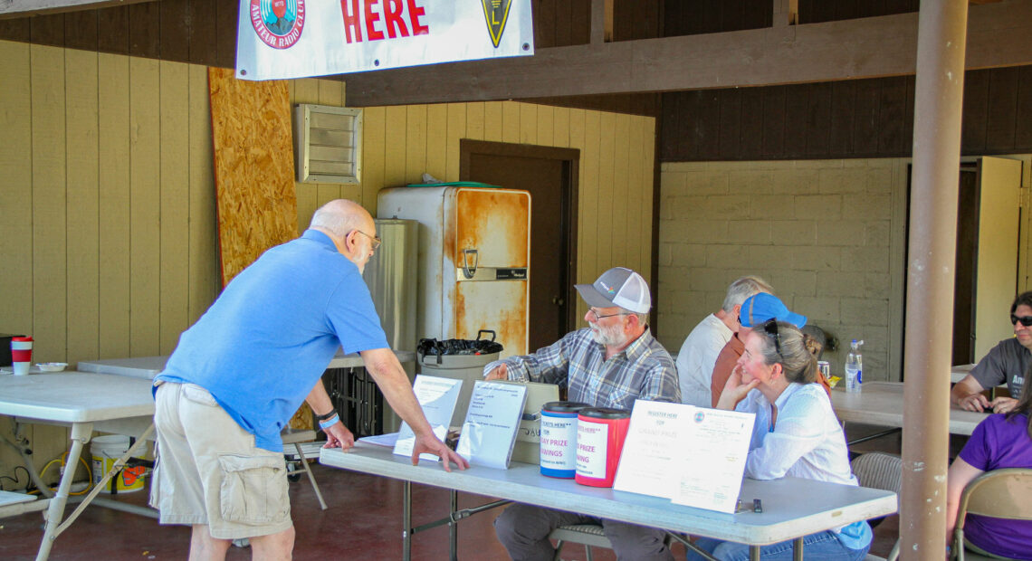 Two people sit at a folding table. A man in a blue shirt and light brown shorts stands in front of the table. A "register here" banner hangs above the table.
