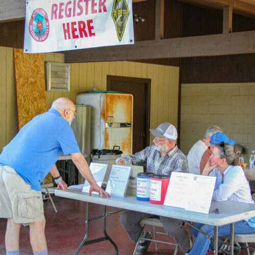 Two people sit at a folding table. A man in a blue shirt and light brown shorts stands in front of the table. A "register here" banner hangs above the table.
