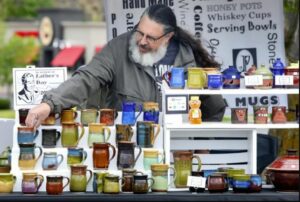 A bearded man wearing glasses and a gray hoodie reaches over a display of brightly colored, handmade mugs. The mugs are displayed on a table, atop of several small shelves.