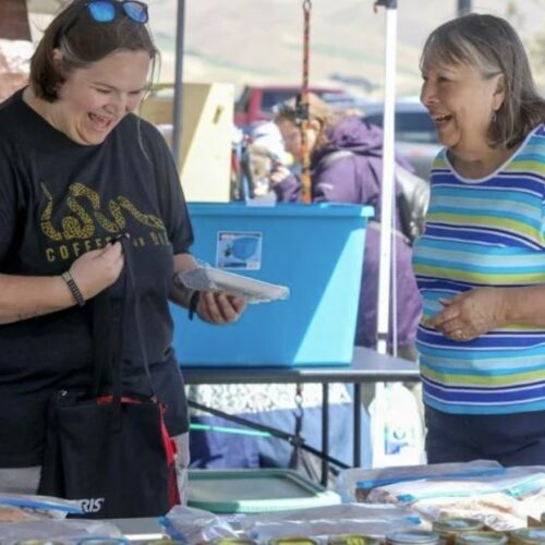 Two women stand in front of a table outside. The woman on the left is wearing a black shirt and is holding a wrapped package. The woman on the right wears a blue striped shirt.