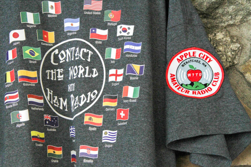 A gray shirt is pictured. The shirt has an Apple City Amateur Radio Club patch on the sleeve. On the front of the shirt, there are many world flags surrounding the phrase "contact the world with ham radio."