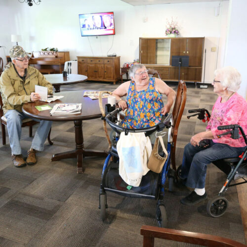Three people are gathered around a circular, brown table having a conversation. A man in brown jacket is seated to the far left. A woman in a blue and pink checked shirt is seated to the right. A woman is seated in a walker next to her.