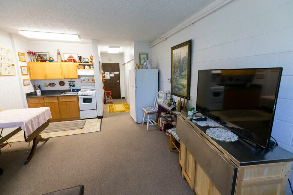 A studio apartment is pictured. A flatscreen TV atop a brown table is pictured to the right. The kitchen, with brown cabinets and a white stove, is pictured in the back.