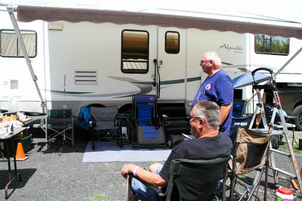 One man in a black shirt sits in a chair. Another man in a blue shirt stands. They are both in front of an RV.