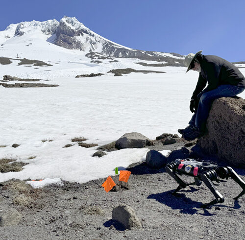 A man dressed in black sits on a gray rock. There is snow behind him. He is looking at a black robot in the shape of a dog. Behind the man and the robot is the summit of Mount Hood.