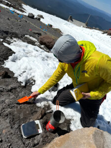 A man in a yellow jacket and gray hat holds a bring orange shovel. In front of him is a rocky area with a camping stove and a black scale. He is kneeling on snow.
