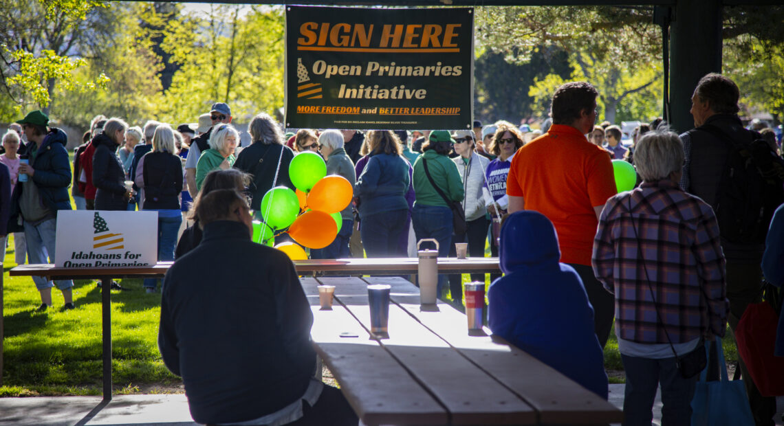 People stand in green grass and sit at a picnic table under a sign that reads "sign here."