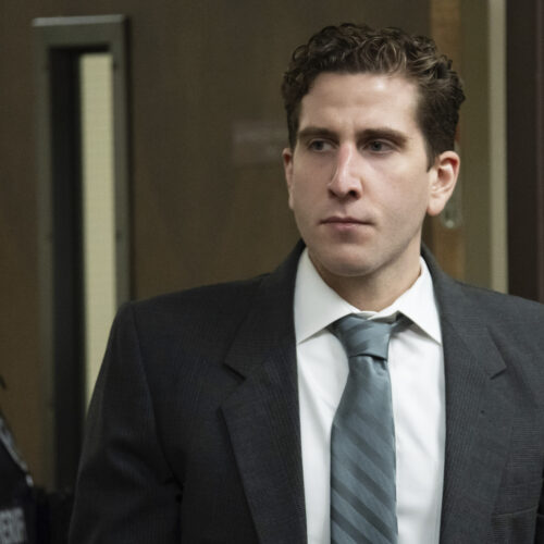 Bryan Kohberger, wearing a gray suit and tie, is escorted into a courtroom in Latah County by a police officer.