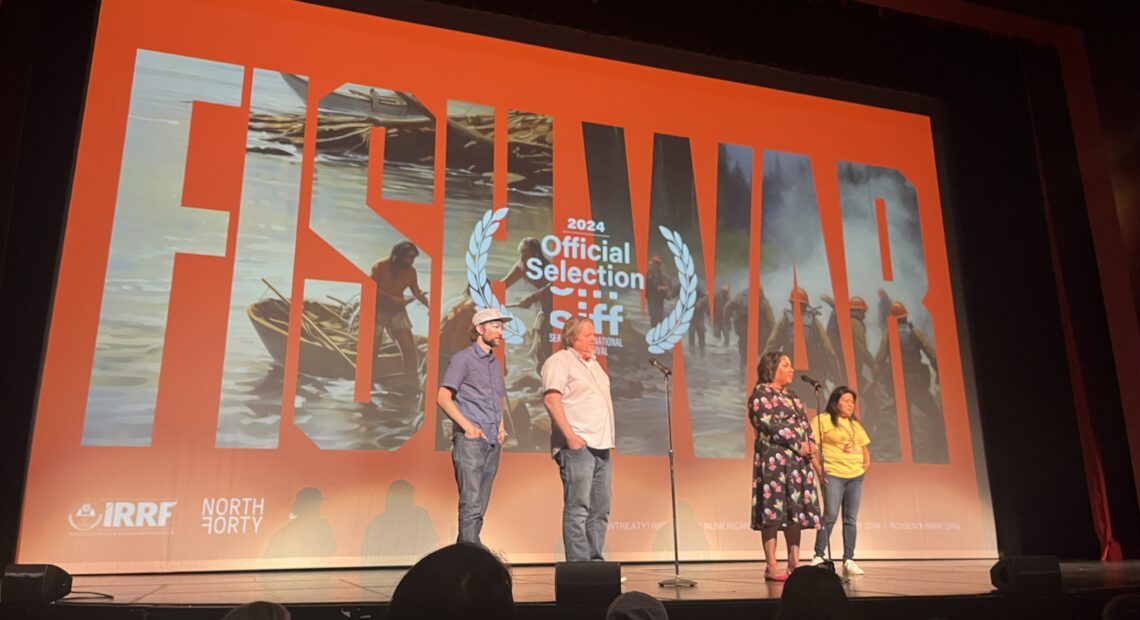 Four people, two men and two women, stand in front of a large screen on a stage. The screen has the words "Fish War" projected on it.