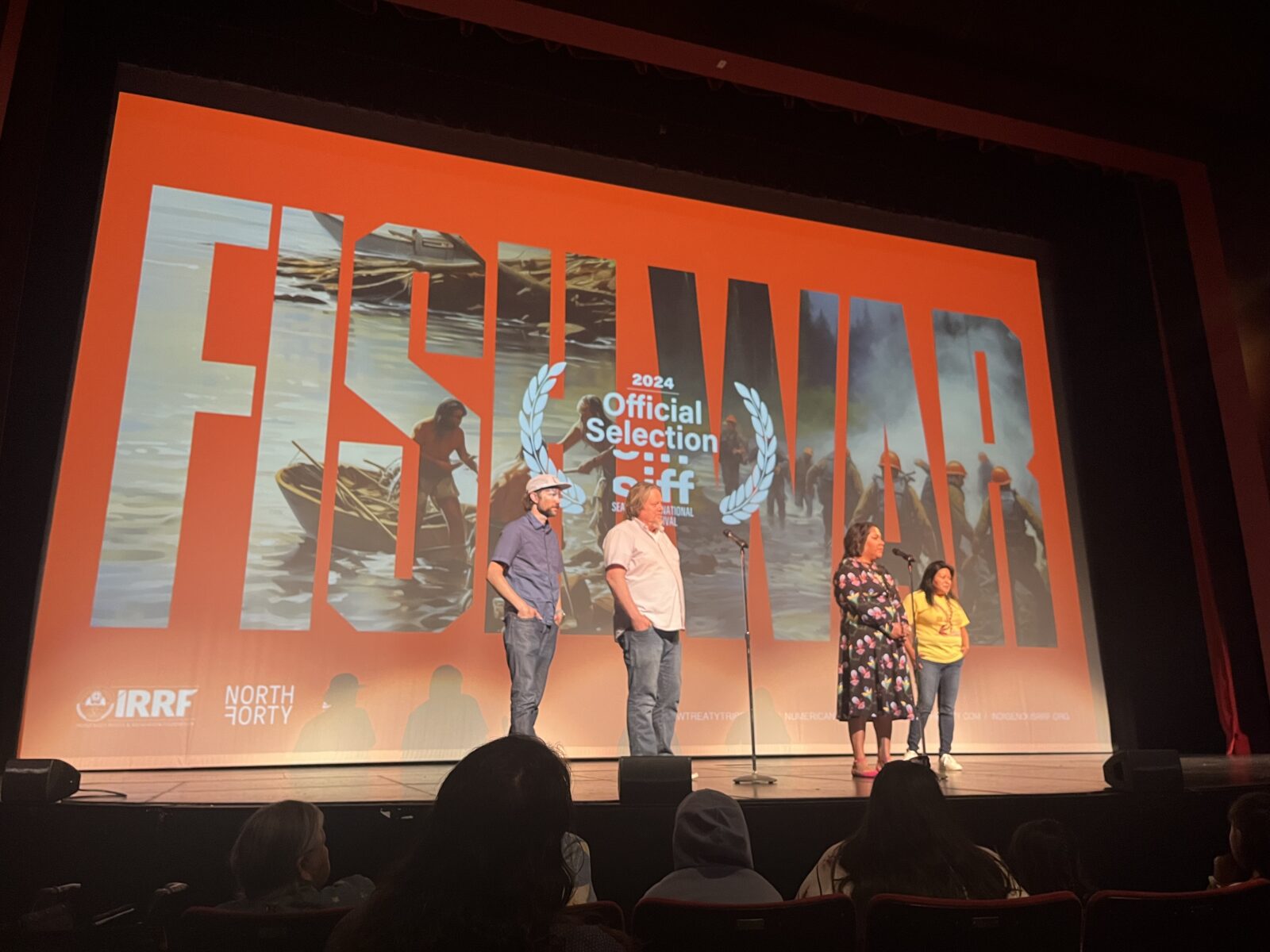 Four people, two men and two women, stand in front of a large screen on a stage. The screen has the words "Fish War" projected on it.