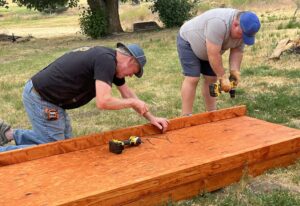 Two men are crouched over a wooden wheelchair ramp. They are using tools to attach curbing to the edge of the ramp.