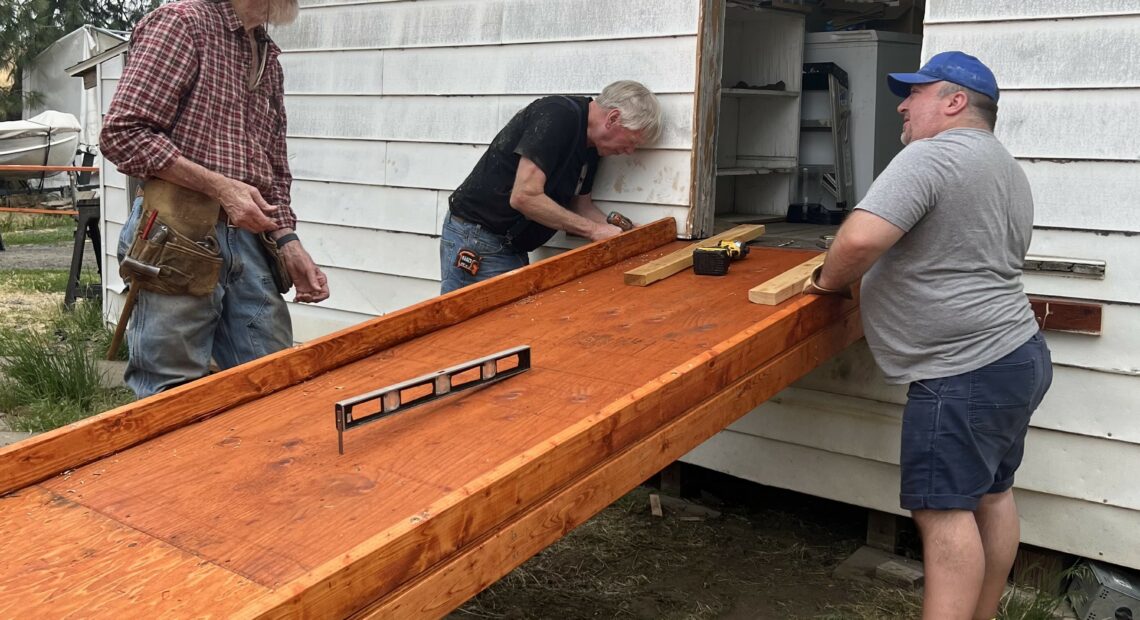 Three men are gathered around the back door of a house. They are working on attaching a wooden wheelchair ramp to the house.