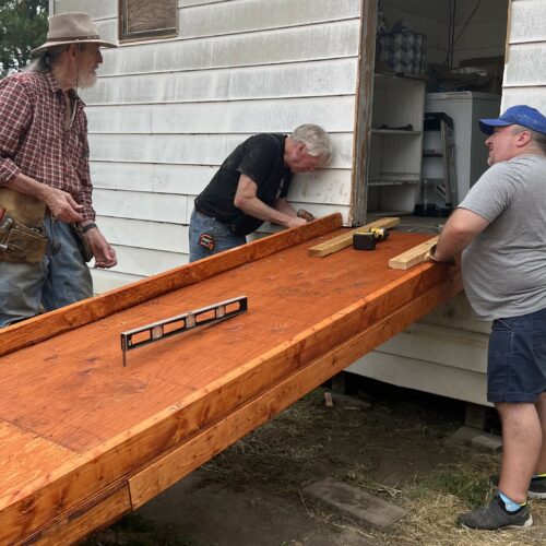 Three men are gathered around the back door of a house. They are working on attaching a wooden wheelchair ramp to the house.