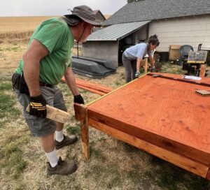 A man in a green shirt and brown hat, and a woman in a light blue shirt, work on a wooden wheelchair ramp. They are making sure the ramp is at the correct grade.