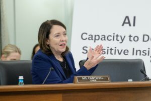 U.S. Senator Maria Cantwell talks into a microphone at a brown wooden podium about AI while wearing a dark blue blazer.