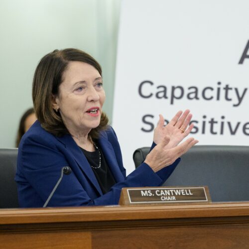 U.S. Senator Maria Cantwell talks into a microphone at a brown wooden podium about AI while wearing a dark blue blazer.