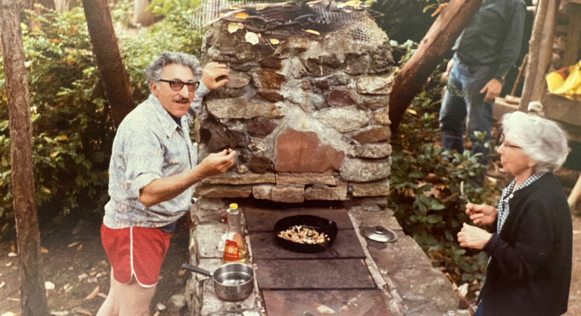 Two people are pictured outdoors frying clams in a cast iron skillet. A man is pictured to the left of the skillet, and a woman is to his right.