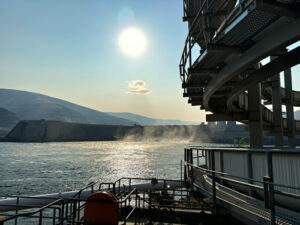 A swirly concrete structure is in front of the Columbia River. The sun is high in the sky. In the background is a portion of Lower Granite Dam.