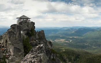 A small house with glass windows surrounding the entire outside sits on top of spiky gray rocks. In the distance are green hills all the way to the horizon. There are gray-white clouds in the sky.