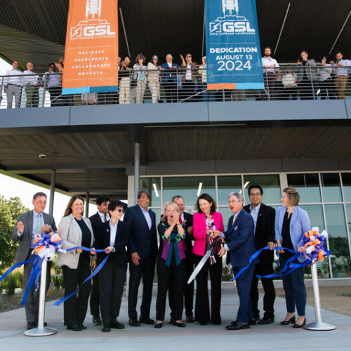 A group of people in suits stand in front of a building, where people are on a balcony. One woman in pink is holding giant scissors to cut a ribbon.