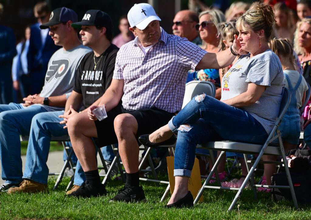 Kaylee Goncalves' parents sit on silver folding chairs on a green lawn. Steve wipes a tear from Kristi's cheek. 