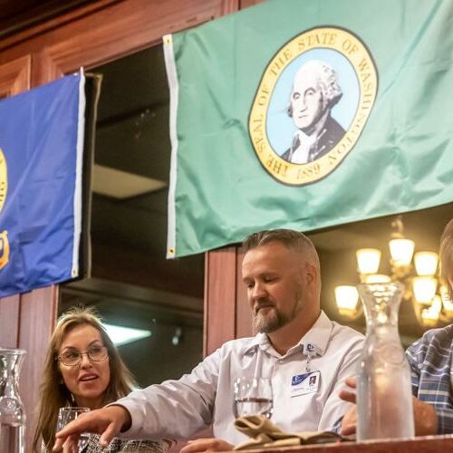 A woman and two men sit at a table with water glasses in front of them. The state flags of Idaho and Washington are behind them.