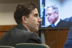 A photo shows the side profile of Bryan Kohberger, who has pale skin and dark hair. A video screen with the gray-haird Judge John Judge on display is in the background.