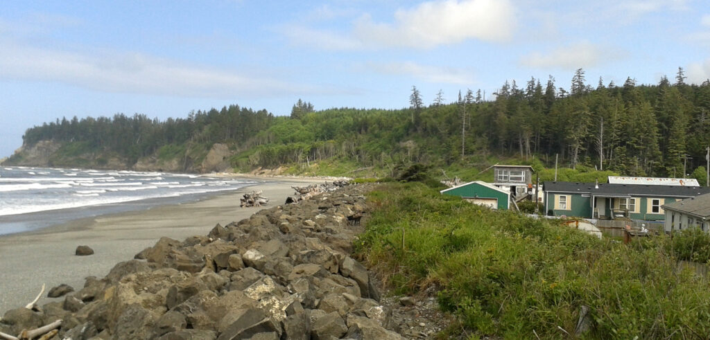 Houses, one painted dark green, are visible near the beach of the Pacific Ocean with waves washing on shore dangerously close to homes. Green mountains are in the background.