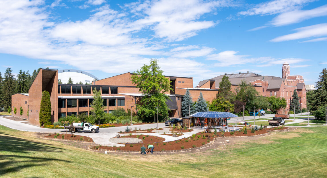 Framed by a blue sky with white clouds and a green field, a round black structure surrounded by brown dirt plots and winding concrete paths sits across from a brick building on the University of Idaho campus.