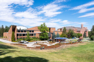 Framed by a blue sky with white clouds and a green field, a round black structure surrounded by brown dirt plots and winding concrete paths sits across from a brick building on the University of Idaho campus.