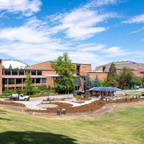 Framed by a blue sky with white clouds and a green field, a round black structure surrounded by brown dirt plots and winding concrete paths sits across from a brick building on the University of Idaho campus.