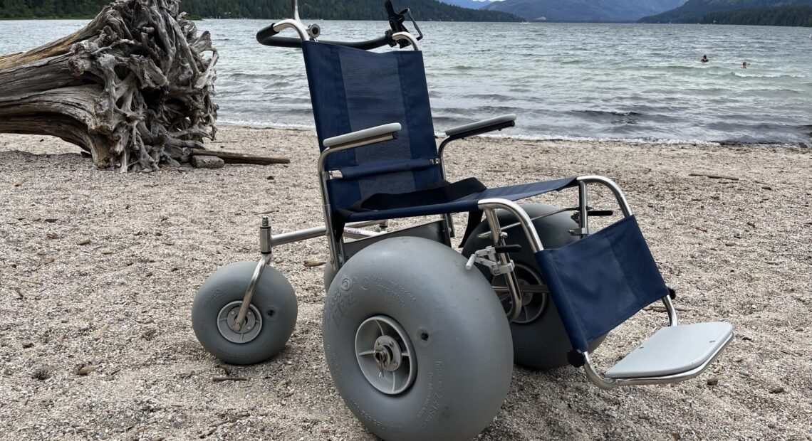 A blue beach wheelchair, with three large gray tires, sits on a beach at Lake Wenatchee.