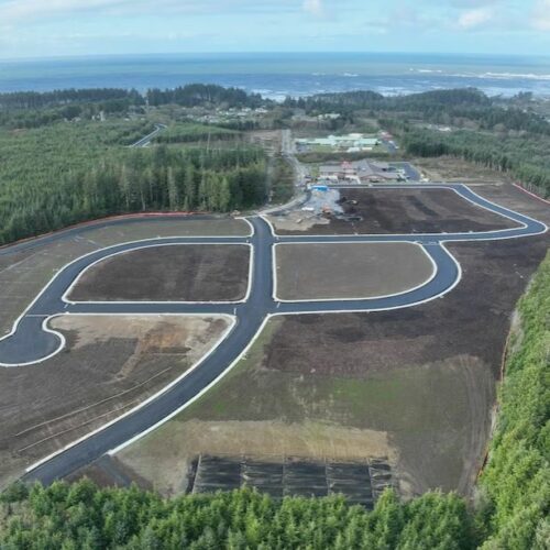 An aerial photo shows six new housing plots with a black paved road surrounded by green trees.