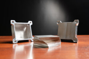 A stack of silver squares sit on top of a wooden table. On either side of the sqaures are two sides of a metal box. The background is black.