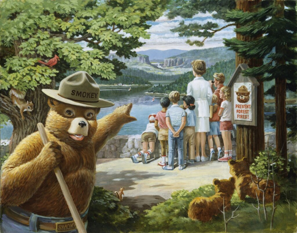 Smokey bear points to a group of people and children looking out over a beautiful lake framed by mountains and trees. A sign hanging on a nearby tree reads, "Prevent Forest Fires."