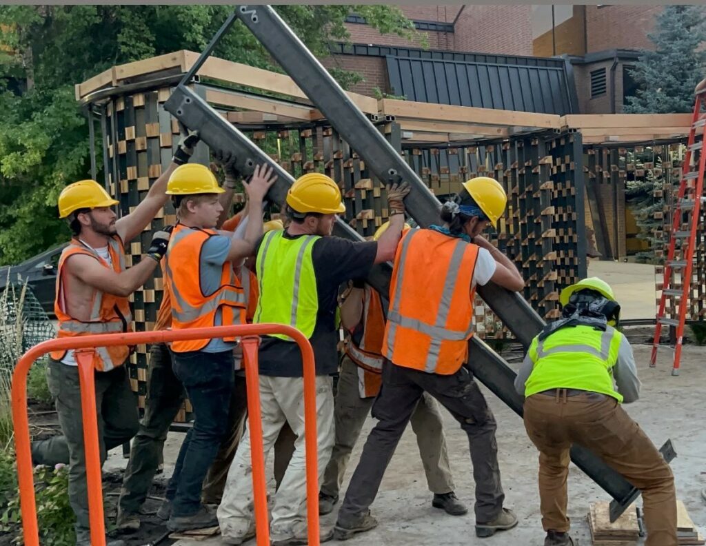 Five young men in yellow hard hats and safety vests move a beam into place on a construction site. 