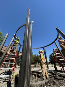 Students in yellow hard hats stand around a circular metal structure on ladders beneath a blue sky. 