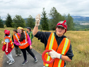 Four girls are in orange vests. One girl in the foreground haas her arm in the air, holding onto a string. They are walking on a cement path that's surrounded by greenish, brownish grass. Evergreen trees are in the background. They sky is cloudy.