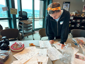 A girl in a black sweatshirt and yellow bandana is standing over an off-white table. She's looking down at white papers spread all over the table.