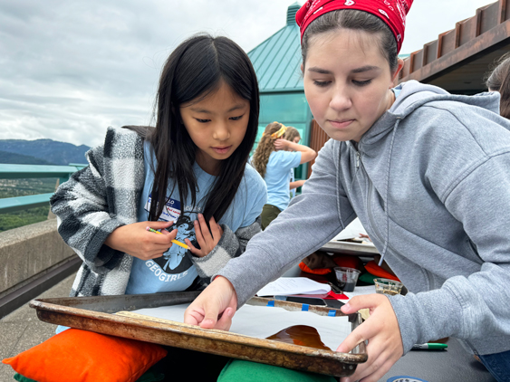 One girl in a blue plaid shirt and another girl in a red bandana and grey shirt are looking at a cookie sheet with brown goop running down it. The sky is cloudy.