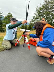 A girl in a blue sweatshirt and orange vest kneels down on grey cement. They are holding long black poles next to an orange tripod with a white disc on top. A woman in a blue shirt with green sleeves holds another long black pole in front of the girl.