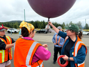 Three girls in orange vests stand around a woman in an orange vest and a green cap. The woman is holding onto a large purple balloon.