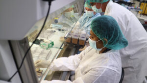 Three scientists are in white lab coats with blue hairnets and face masks. They are holding what appears to be dirt in small containers.