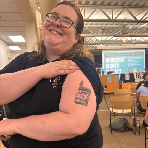 Ann Dorn, who serves on the steering committee of Tacoma for All, poses at the workers' rights event with the tattoo she got after the group's initiative for a tenant bill of rights passed in 2023. (Credit: Lauren Gallup / NWPB)