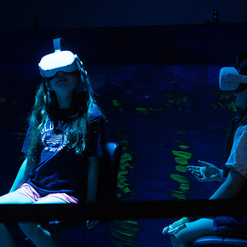 Two girls with virtual reality headsets are in a dark room. One of them is looking up towards the ceiling.